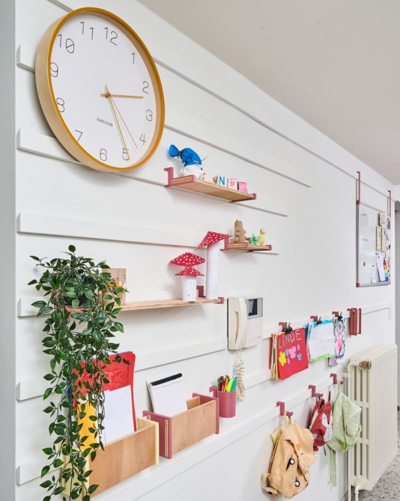 WandOrde modulair wandsysteem in de leefruimte met legplanken, haken, documenthouder, kindertekeningen en decoratie.