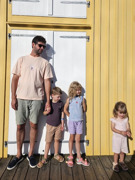 Vader met drie jonge kinderen buiten aan een houten wand – gezinsleven en dagelijkse organisatie, inspiratie voor WandOrde wandsystemen.